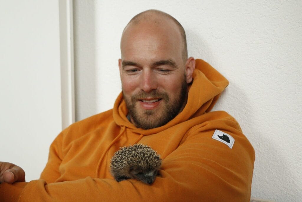Ein Mann mit Glatze und Bart trägt einen orangefarbenen Hoodie. Er hält einen kleinen Igel auf seinem Arm und lächelt dabei in Richtung des Tiers.